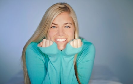 Young blond woman smiling with head in hands and perfect white teeth, gray backdrop