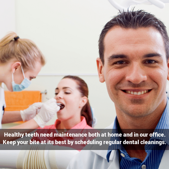 Dentist looking at the camera while woman gets dental cleaning in the background