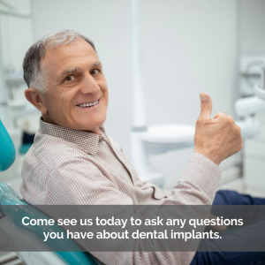 An elderly man gets dental implants. He sits in a dental chair giving the thumbs up with a smile.