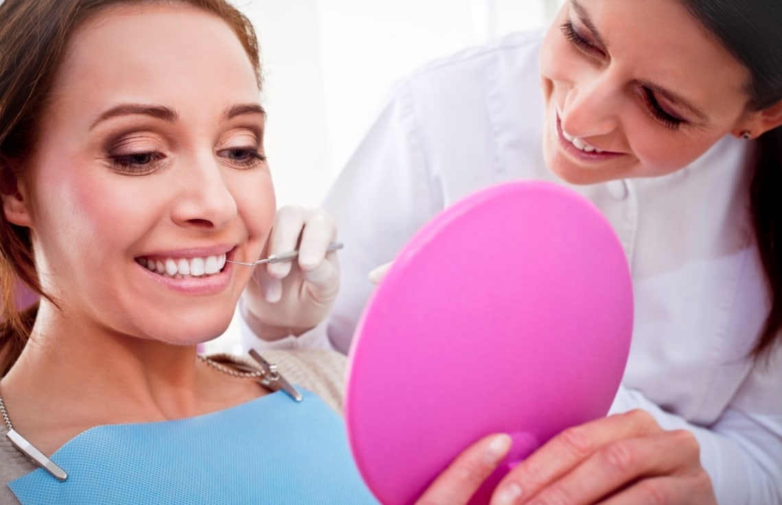 Female dentist with dental tool, woman in dental chair smiling and looking at hand mirror
